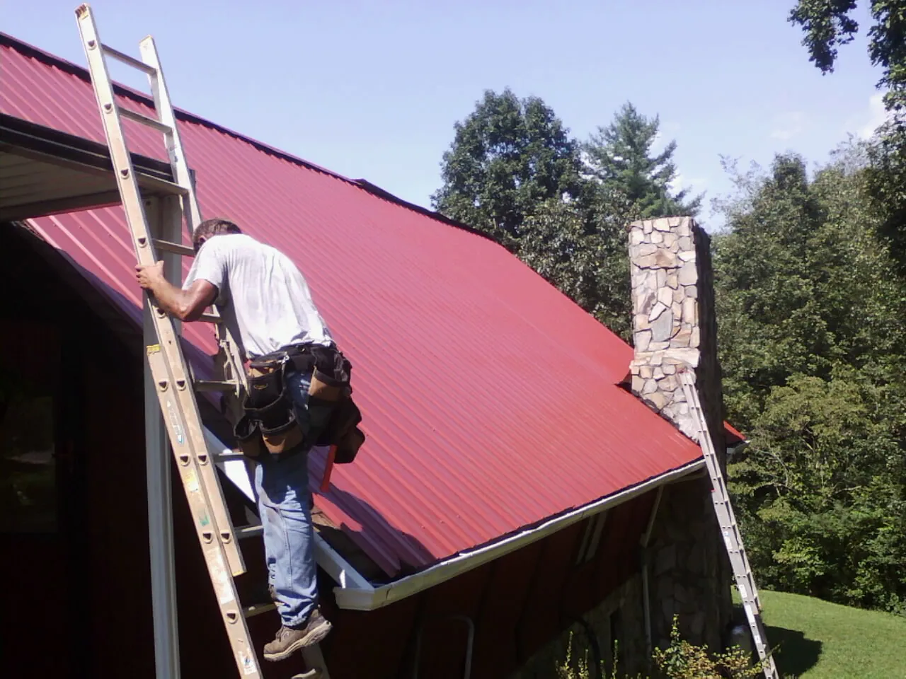 Roofer on a ladder beside a red metal roof and stone chimney — Soffit & Fascia Repair by Roofing Brothers in Corydon